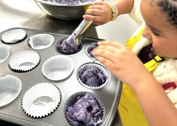 A young girl is baking purple cupcakes, carefully placing the batter in a muffin tin.